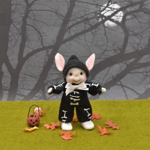 May include: A small, white, felt bunny wearing a black and white skeleton costume and a black hat. The bunny is holding a small orange pumpkin bucket and is standing on a green surface with red and orange leaves scattered around it. The background is a dark gray silhouette of trees.