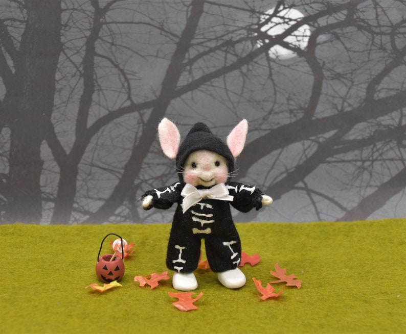 May include: A small, white, felt bunny wearing a black and white skeleton costume and a black hat. The bunny is holding a small orange pumpkin bucket and is standing on a green surface with red and orange leaves scattered around it. The background is a dark gray silhouette of trees.