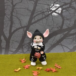 May include: A small, white, felt mouse wearing a black skeleton costume and a black witch hat, holding a small orange pumpkin bucket. The mouse is standing on a green grassy surface with scattered autumn leaves. The background is a silhouette of bare tree branches against a dark sky with a full moon.