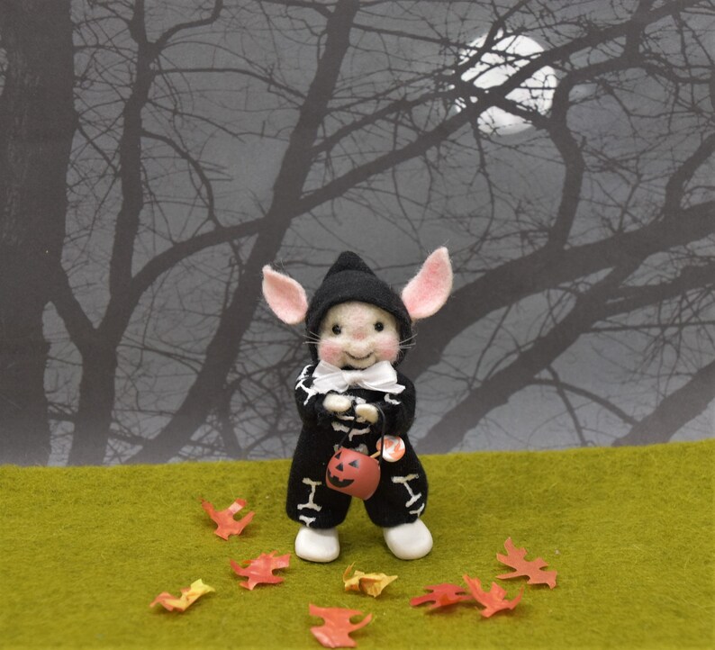 May include: A small, white, felt mouse wearing a black skeleton costume and a black witch hat, holding a small orange pumpkin bucket. The mouse is standing on a green grassy surface with scattered autumn leaves. The background is a silhouette of bare tree branches against a dark sky with a full moon.