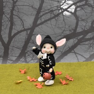 May include: A small, felt mouse wearing a black and white skeleton costume and a black hat. The mouse is holding a small orange jack-o'-lantern bucket. The mouse is standing on a green surface with orange leaves scattered around it. The background is a blurry image of a dark forest with a full moon.