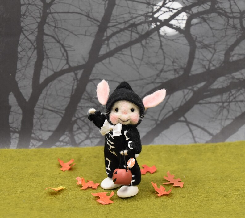 May include: A small, felt mouse wearing a black and white skeleton costume and a black hat. The mouse is holding a small orange jack-o'-lantern bucket. The mouse is standing on a green surface with orange leaves scattered around it. The background is a blurry image of a dark forest with a full moon.