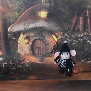 May include: A small, felt mouse dressed in a black skeleton costume and a black witch hat holds a small orange pumpkin. The mouse is standing in front of a miniature mushroom house with a lit window and a wooden door.