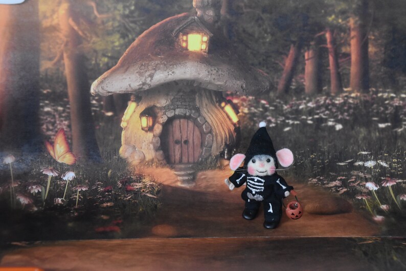 May include: A small, felt mouse dressed in a black skeleton costume and a black witch hat holds a small orange pumpkin. The mouse is standing in front of a miniature mushroom house with a lit window and a wooden door.