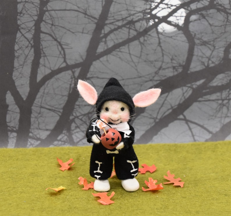 May include: A small, white rabbit wearing a black and white skeleton costume and holding a small orange jack-o'-lantern. The rabbit is standing on a green grassy surface with fallen autumn leaves.