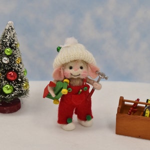 May include: A small, white, felt mouse wearing a white knit hat and red overalls is holding a toy hammer and a green toy truck. The mouse is standing in front of a small Christmas tree and a wooden toolbox with tools.