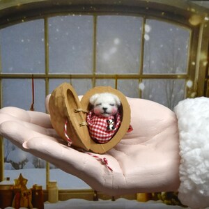 May include: A miniature white puppy figurine inside a walnut shell, on a red and white checked blanket. The shell is held open, revealing the puppy. The background shows a snowy window scene.