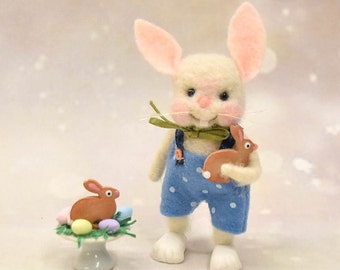 May include: A white felt bunny wearing blue overalls with white polka dots holds a brown felt bunny. The bunny is holding a green ribbon around its neck. The bunny is standing on a white surface with a small cake stand with a brown felt bunny and pastel colored eggs on it.