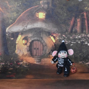 May include: A small, felt mouse dressed in a black skeleton costume and a black witch hat holds a small orange pumpkin. The mouse is standing in front of a miniature mushroom house with a lit window and a wooden door.