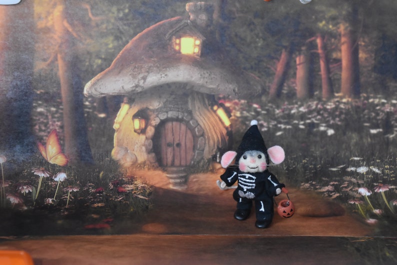 May include: A small, felt mouse dressed in a black skeleton costume and a black witch hat holds a small orange pumpkin. The mouse is standing in front of a miniature mushroom house with a lit window and a wooden door.