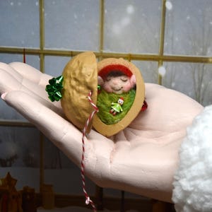 May include: A charming Christmas decoration showing a miniature elf inside a walnut shell. The elf is dressed in a red hat and green outfit, resting on green felt. The walnut is decorated with a green bow and a red and white string.