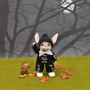 May include: A small, white, felt bunny wearing a black skeleton costume and a white bow tie. The bunny is holding a small orange pumpkin bucket with candy. The bunny is standing on a green surface with orange leaves scattered around it. The background is a blurry image of a dark forest.
