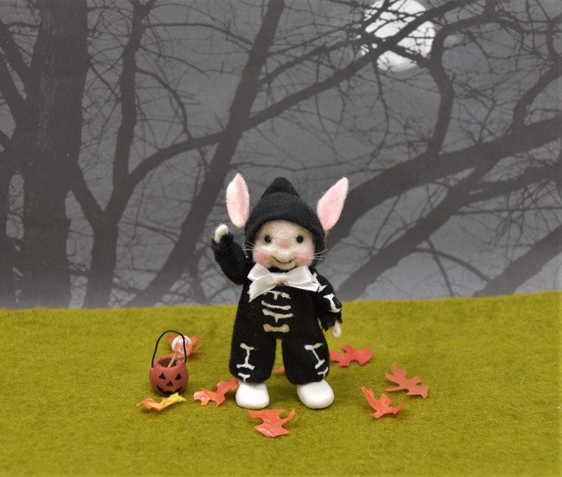 May include: A small, white, felt bunny wearing a black skeleton costume and a white bow tie. The bunny is holding a small orange pumpkin bucket with candy. The bunny is standing on a green surface with orange leaves scattered around it. The background is a blurry image of a dark forest.