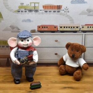 May include: A needle-felted mouse figurine wearing a blue and white striped engineer cap and matching overalls, holding a miniature toy train engine. Beside the mouse sits a brown plush teddy bear wearing a white shirt, with a small train car nearby.