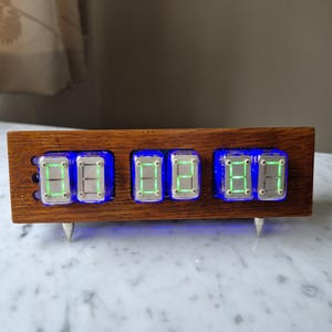 May include: A digital clock with a wooden frame displays the time 08:08. The clock features blue neon lighting around the digital displays and is supported by two silver metal stands. The numbers are displayed in green.