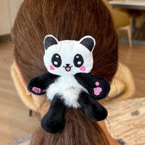 May include: A plush panda hair scrunchie featuring a white and black panda face with pink cheeks, black ears, and black limbs. The center of the accessory is made of white faux fur, while the panda limbs are black with pink heart-shaped paw pads.