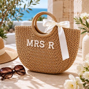 May include: A woven straw tote bag with a rounded shape and a handle. The bag features the embroidered letters "MRS R" and a white satin bow. Tortoiseshell sunglasses and a straw hat are also visible.