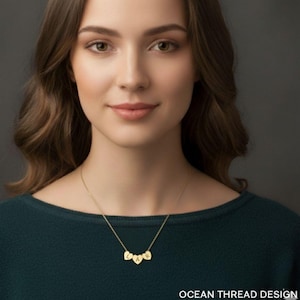 May include: A gold-colored necklace with three heart-shaped pendants, each bearing a single letter. The letters spell out "LAR". The necklace is worn by a person wearing a dark green top. The text "OCEAN THREAD DESIGN" is at the bottom.