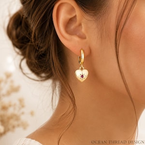 May include: A gold hoop earring featuring a dangling heart-shaped charm. The heart has a polished gold finish and is centred with a small, star-engraved detail containing a tiny, vibrant red gemstone. The text OCEAN THREAD DESIGN is visible.
