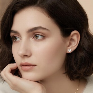 May include: A woman wearing small, silver stud earrings. The earrings feature a heart-shaped design. The woman is wearing a cream-coloured sweater and a gold necklace. The image is well-lit, with a soft focus on the woman's face.