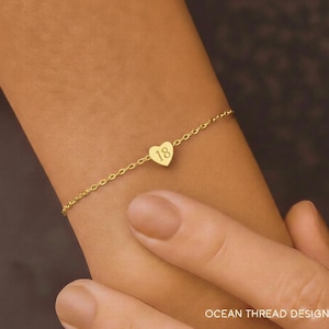Op de afbeelding: Een delicate goudkleurige armband met een hartvormige bedel. De bedel heeft het nummer "18" gegraveerd. De armband zit om een pols. De tekst "OCEAN THREAD DESIGN" staat onderaan de afbeelding.