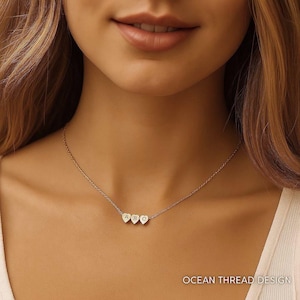 Può includere: Una delicata collana dorata con tre ciondoli a forma di cuore. Ogni ciondolo ha una lettera incisa. La collana è indossata intorno al collo. Il testo "OCEAN THREAD DESIGN" è in basso.