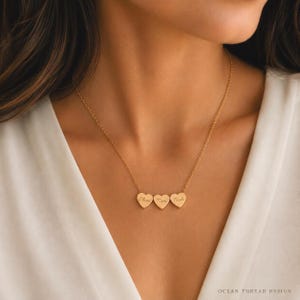 May include: A gold-toned necklace featuring three small heart-shaped pendants suspended from a delicate chain. Each heart is engraved with a name in a cursive script, specifically reading Olivia, Mom, and Noah from left to right.