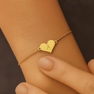 May include: Gold-coloured bracelet with a heart-shaped charm bearing the letter "A". The fine chain bracelet is worn on a wrist, with a hand in the foreground. The bracelet is a piece of jewellery.