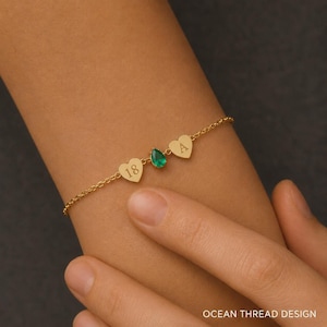 May include: A gold-toned chain bracelet featuring two small heart-shaped charms and a central pear-shaped green gemstone. One heart charm is engraved with the number 18, and the other heart charm is engraved with the capital letter A.