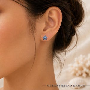 May include: A star-shaped stud earring featuring five marquise-cut blue gemstones arranged in a floral pattern with a small clear stone at the centre, set in a metallic frame. The text "OCEANTHREAD DESIGN" is visible in the bottom right corner.