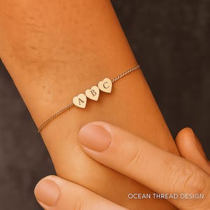 Puede incluir: Una pulsera dorada con tres colgantes en forma de corazón, cada uno grabado con una letra: A, B y C. La pulsera está en una cadena fina. El texto "OCEAN THREAD DESIGN" es visible en la parte inferior derecha.