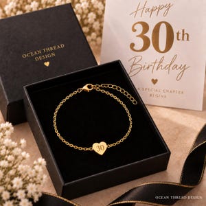 Op de afbeelding: Een goudkleurige kettingarmband met een kleine hartvormige bedel gegraveerd met het getal 30. De armband wordt gepresenteerd in een zwarte geschenkdoos naast een kaart met de tekst Happy 30th Birthday A SPECIAL CHAPTER BEGINS.