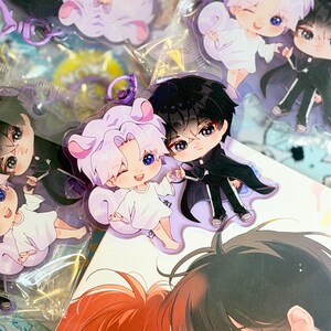May include: Acrylic keychain featuring two chibi-style characters: one with light purple hair and animal ears wearing a white outfit, and one with black hair wearing a black suit. The keychain has a purple plastic clasp and a clear acrylic base.