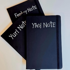 May include: Three black rectangular notebooks with elastic closure bands. Each cover features white hand-lettered text: one reads Femboy Note, another Yuri Note, and the third YAOI NOTE. The notebooks are arranged in a stacked, overlapping formation.