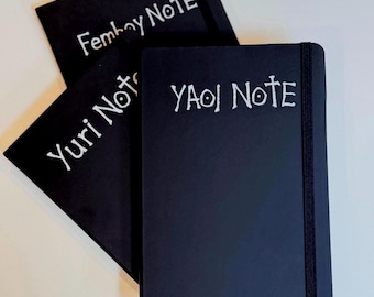Può includere: Tre quaderni rettangolari neri con elastici di chiusura. Ogni copertina presenta un testo bianco scritto a mano: uno riporta Femboy Note, un altro Yuri Note e il terzo YAOI NOTE. I quaderni sono disposti in una formazione sovrapposta.