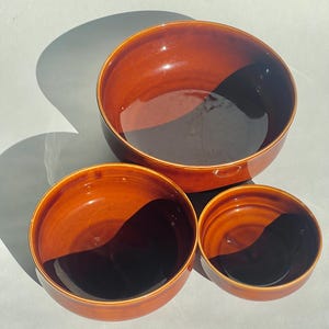 May include: Three glazed ceramic bowls in varying sizes. The bowls are a warm, reddish-brown color with a glossy finish. The bowls are nested together, with the largest bowl at the top and the smallest at the bottom.