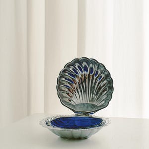 May include: A silver-colored, shell-shaped trinket box with a hinged lid, open to reveal a deep blue interior. The box sits on a white surface, with a white curtain in the background.