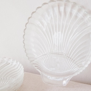 Puede incluir: Plato de servir de vidrio transparente en forma de concha, con borde festoneado. El plato tiene un diseño en forma de abanico con líneas verticales. También se ve otro plato más pequeño y similar.