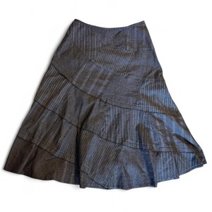 Puede incluir: Una falda de corte en A de color gris oscuro construida con múltiples paneles de tela. La falda presenta un diseño de patchwork que alterna rayas verticales y un sutil patrón de cuadros, creando un aspecto texturizado con dobladillo acampanado.