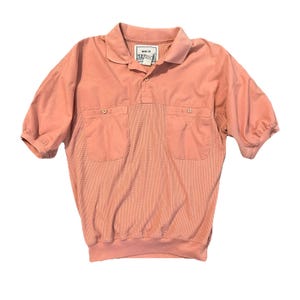 May include: A peach-colored short-sleeve polo shirt with a collared neckline and two front pockets. The shirt has a textured, checkered pattern on the lower half and a smooth fabric on the upper half. The shirt has a button-up placket and a label that reads "MEN'S BY HUK".