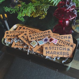 May include: A collection of metal stitch markers attached to rectangular kraft paper cards labeled STITCH MARKERS with star icons. The markers feature small square crochet motifs in various colors, silver-toned charms, and beads in moon and star shapes.