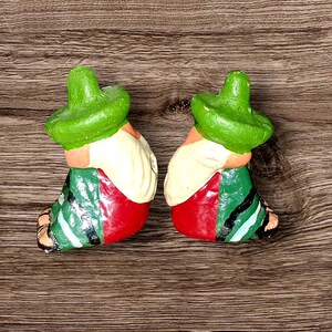 May include: Two ceramic figurines of a seated man wearing a green sombrero, a cream-colored shirt, red pants, and a green and black striped sash. The figurines are placed on a wooden surface.