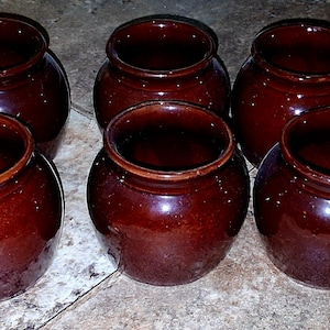 May include: Six glazed, brown ceramic pots with a rounded shape and wide openings. The pots are arranged in a group, showcasing their glossy finish and rustic appeal. The pots are suitable for kitchen or home decor.