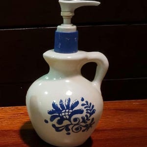 May include: White ceramic soap dispenser with a blue floral design. The dispenser has a blue pump and a handle. The dispenser is on a wooden surface.
