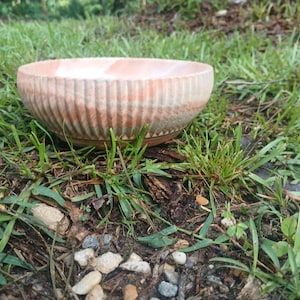 Puede incluir: Un cuenco redondo de cerámica hecho a mano con un exterior acanalado. El cuenco tiene una combinación de colores rosa claro y blanco. El cuenco está sobre un lecho de hierba verde y pequeñas rocas.