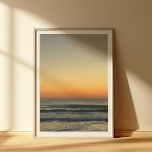 Può includere: Stampa incorniciata di un paesaggio marino al tramonto. Il cielo passa da un blu pallido a vibranti tonalità arancioni e gialle. Dolci onde si infrangono sulla riva, riflettendo i colori caldi del sole al tramonto. L'opera è esposta in una cornice bianca.