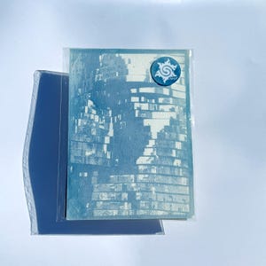 May include: A rectangular cyanotype print on textured paper featuring a monochromatic blue and white abstract architectural pattern of stacked brick-like shapes. A small circular blue sticker with a white stylized S logo is attached.