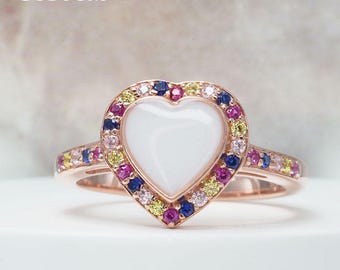 Puede incluir: Un anillo de oro rosa con un diseño en forma de corazón. El corazón central es blanco, rodeado por un halo de piedras preciosas de colores. La banda también presenta piedras preciosas. La palabra "CUSTOM" está en la parte superior.