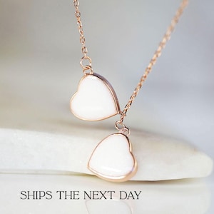 May include: A delicate rose gold necklace with two white heart pendants. The necklace is on a white surface with the text "SHIPS THE NEXT DAY".