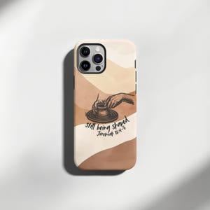 Op de afbeelding: Een telefoonhoesje met een beige, bruin en wit ontwerp. De hoes heeft een illustratie van handen die aardewerk vormen, met de tekst "still being shaped Jeremiah 18:4-6" in een handschrift. De hoes heeft een glanzende afwerking.